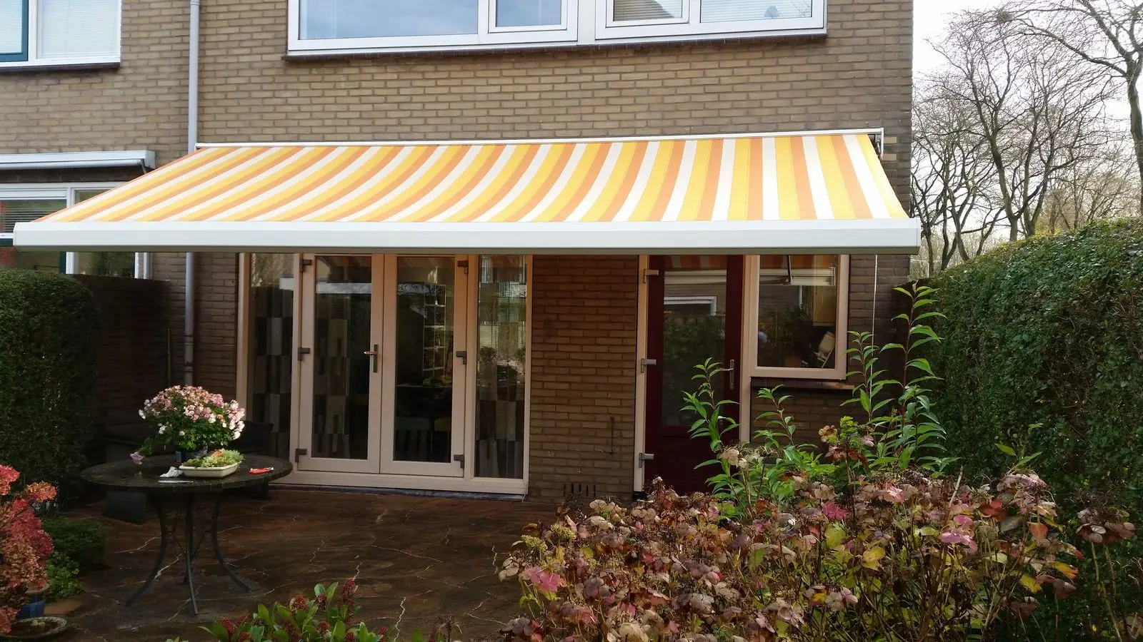 Knikarmscherm met geel-wit gestreept doek boven het terras van een bakstenen woning