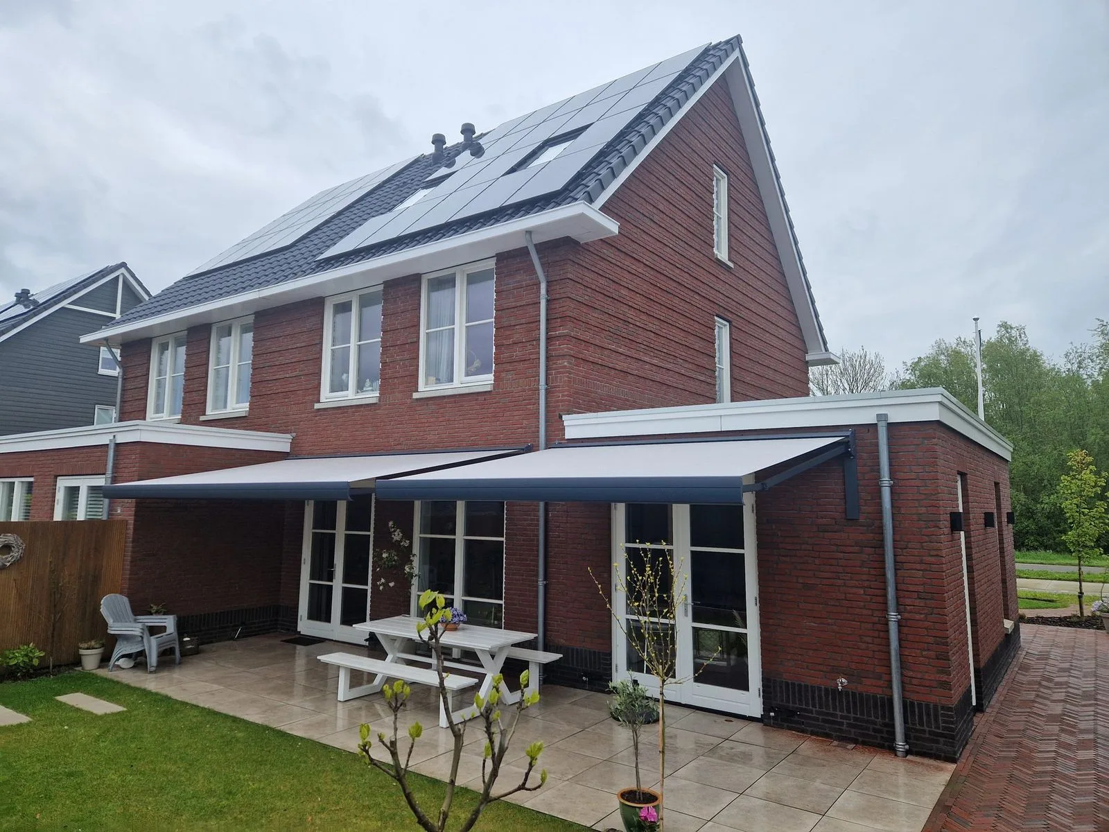Twee antracietgrijze knikarmschermen boven het terras van een bakstenen woning met zonnepanelen