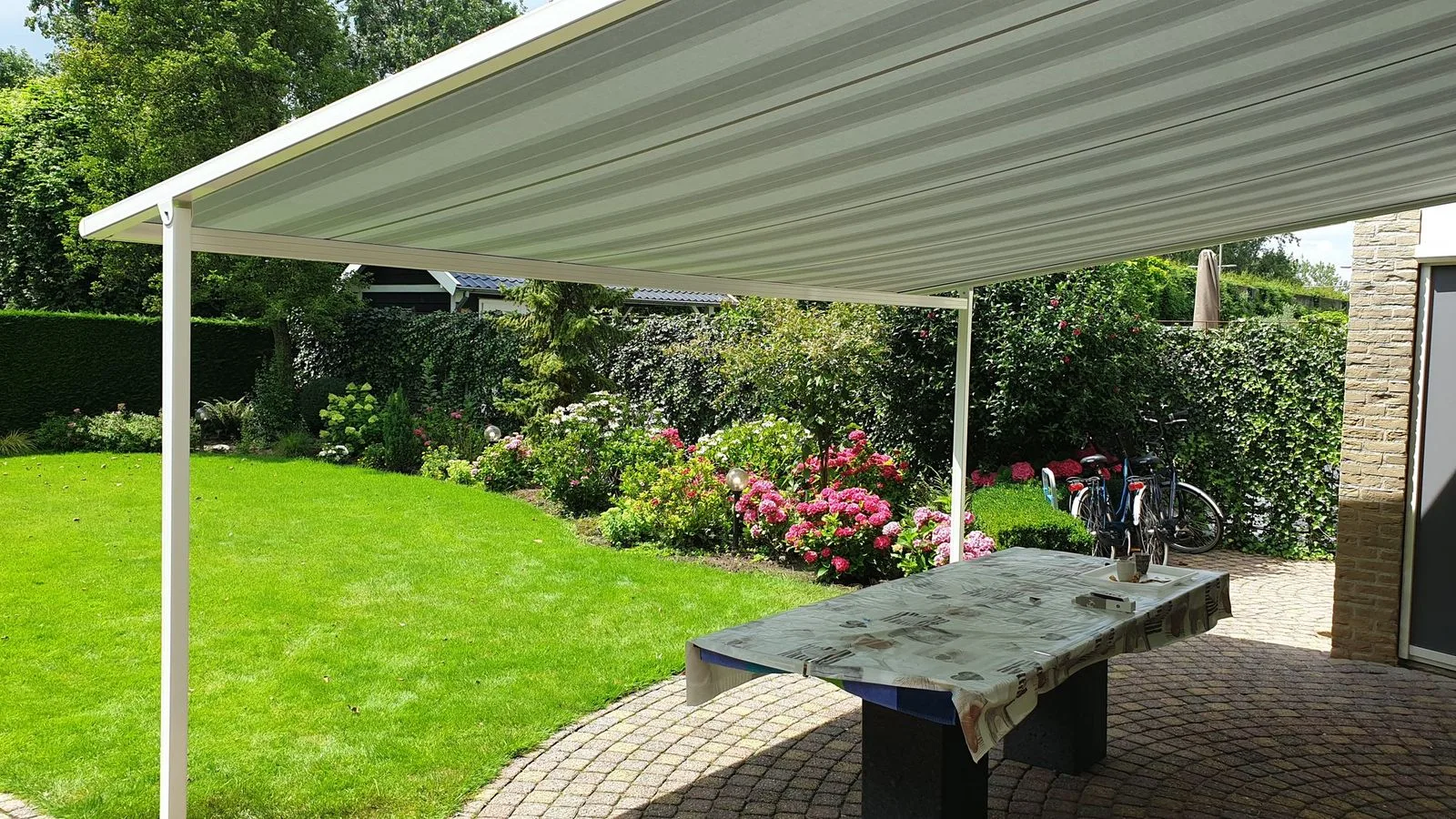 Witte pergolazonwering op palen boven een tuintafel in een groene tuin