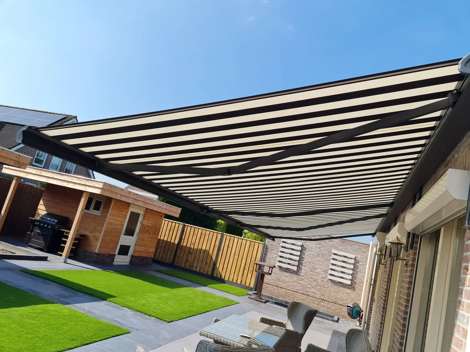Knikarmscherm met grijs gestreept doek, van onderen gezien, boven een terras met kunstgras