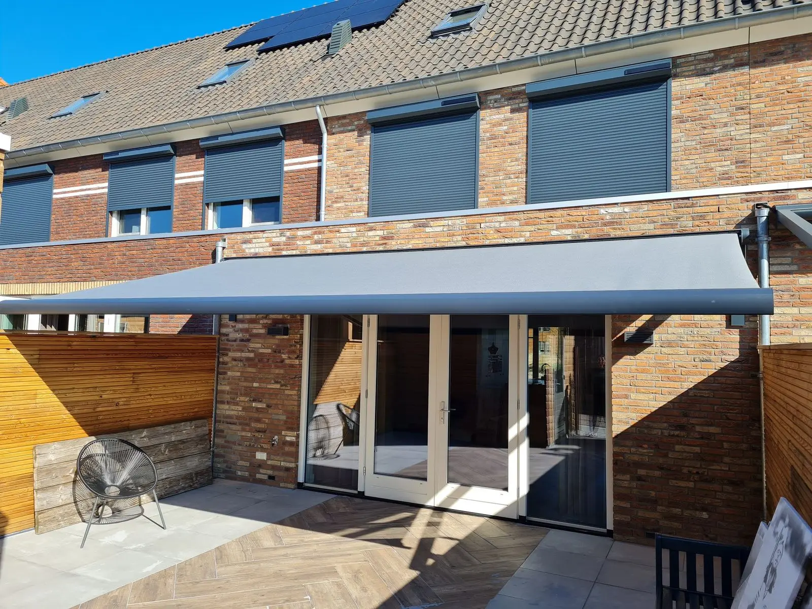 Grijs knikarmscherm boven de tuindeuren van een rijtjeswoning met rolluiken op de verdieping