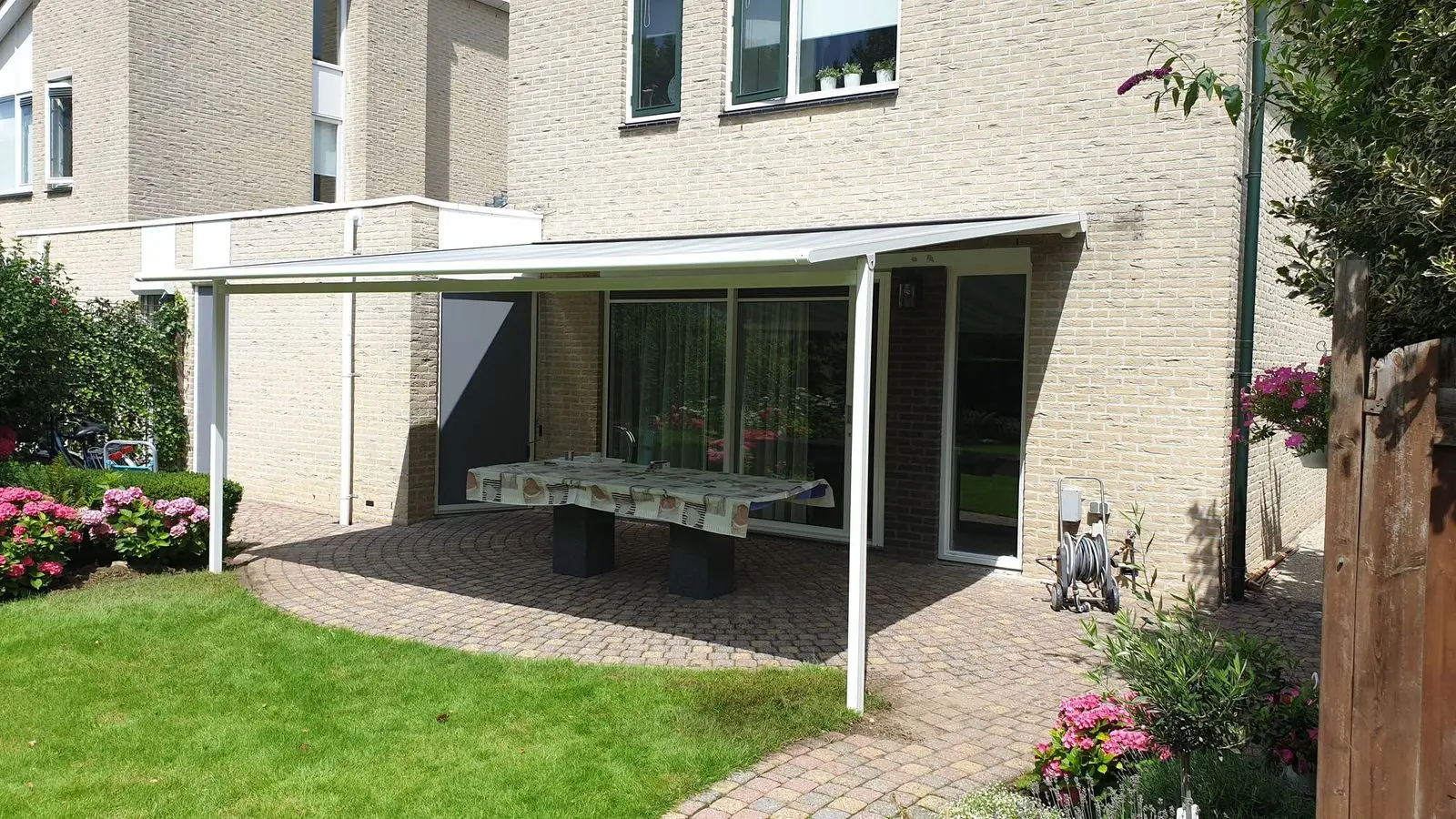 Witte pergolazonwering boven een terras aan een gele bakstenen woning