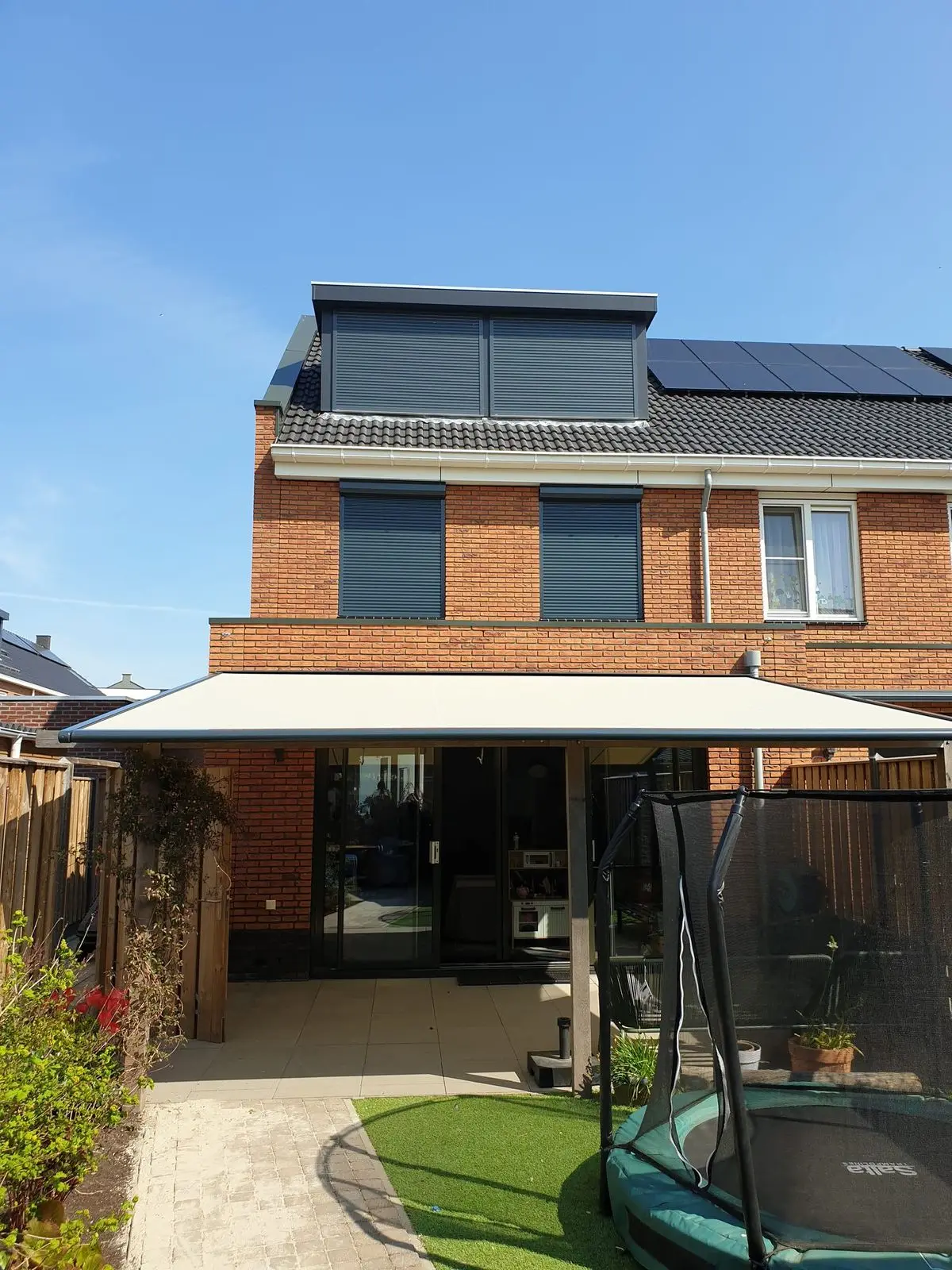 Zonwering over het dak van een aanbouw achter een bakstenen woning, gezien vanuit de tuin